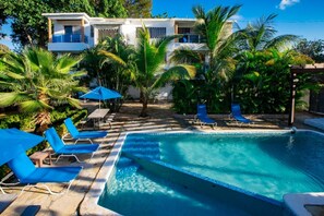 Un espace piscine avec des chaises longues bleues et des parasols, entouré de palmiers et d’un bâtiment blanc doté de balcons.