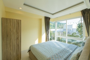 A bedroom with a large window, a bed with patterned bedding, and a wooden wardrobe.
