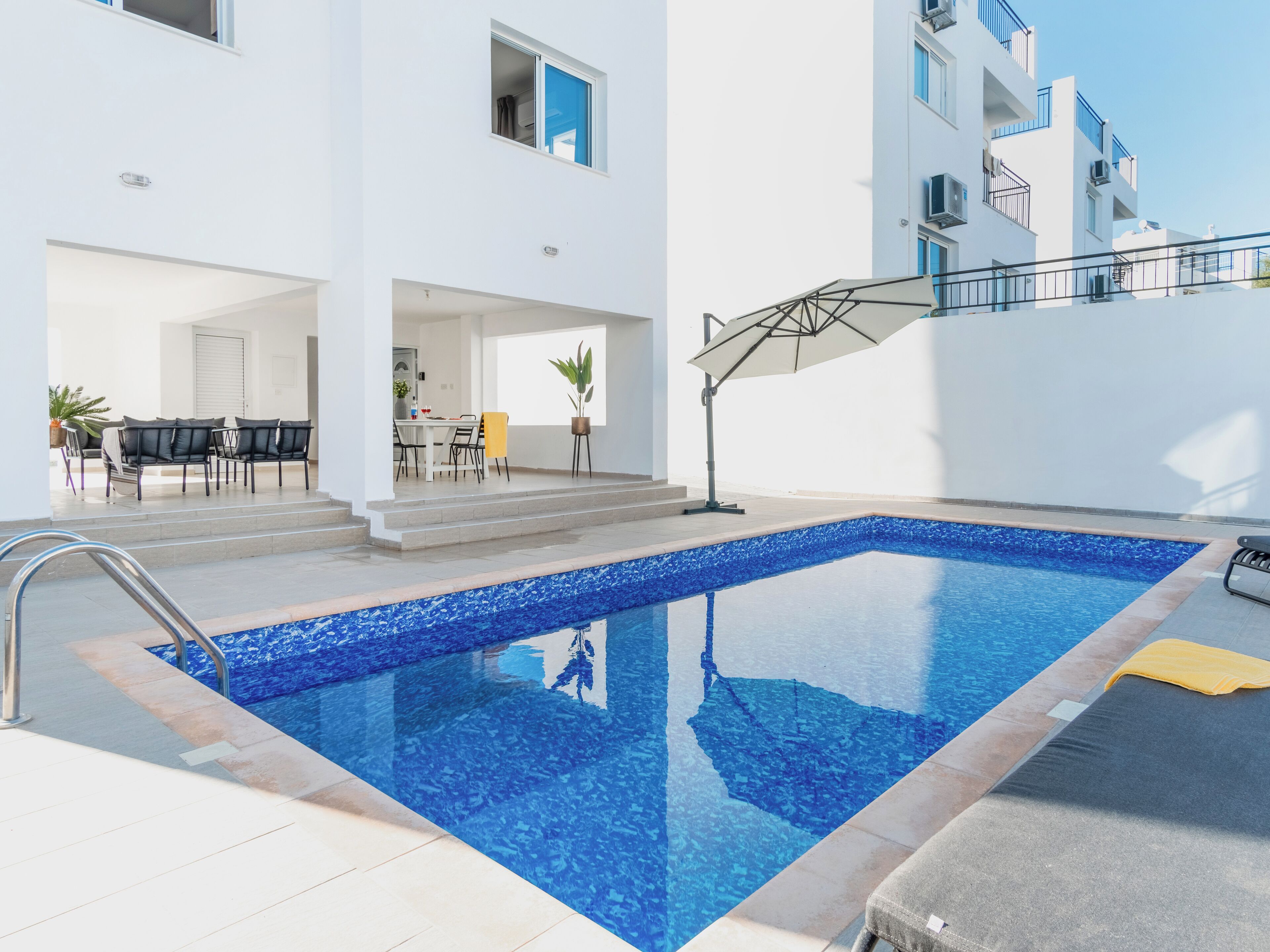 Una piscina exterior moderna con agua azul cristalina, rodeada por un edificio blanco con balcones y un patio cubierto.