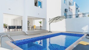 A modern outdoor swimming pool with clear blue water, surrounded by a white building with balconies and a covered patio area. - Sanders White Mountains - Charming Villa With Pool (Tremithousa)