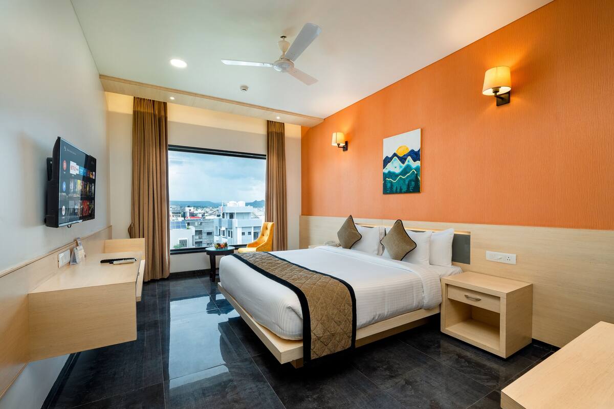 Ein modernes Hotelzimmer mit einem großen Bett, einem Schreibtisch, einem Sessel, einem Fernseher und Blick auf die Stadt.