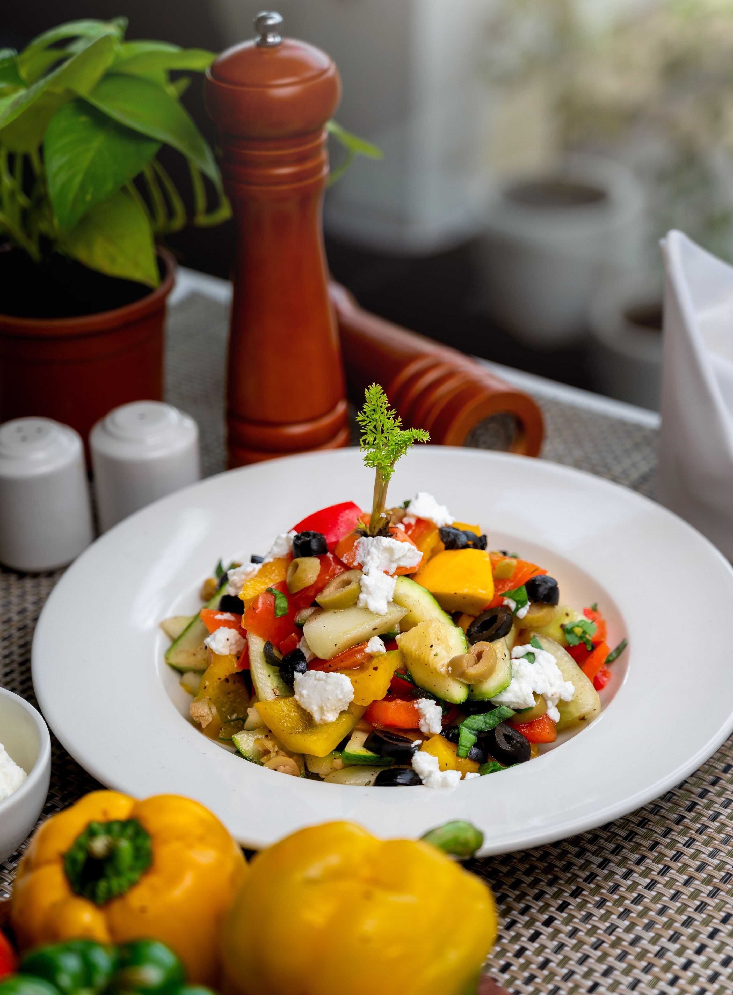 A plate of colorful vegetable salad with feta cheese on a white plate, surrounded by yellow and green bell peppers, and a pepper grinder in the background.