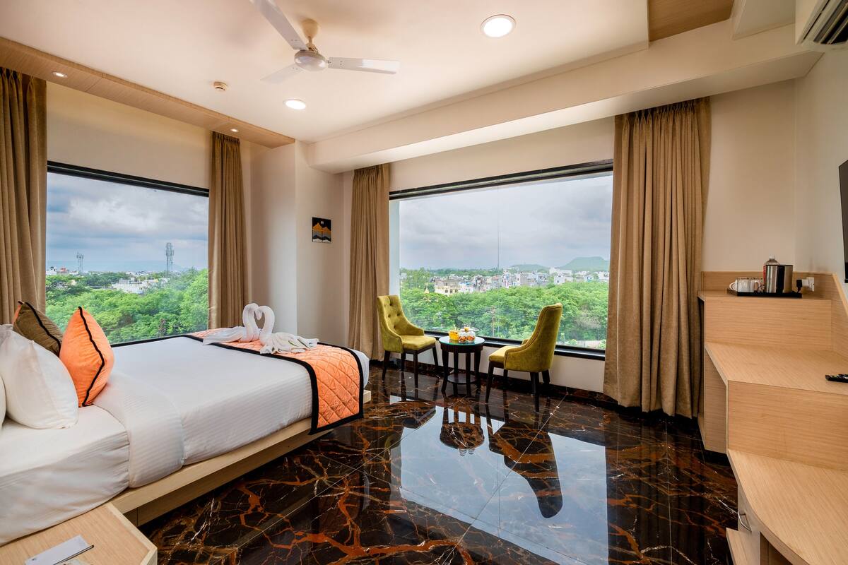 Ein Hotelzimmer mit einem Bett, zwei Stühlen, einem kleinen Tisch und großen Fenstern mit Blick auf die Stadt.