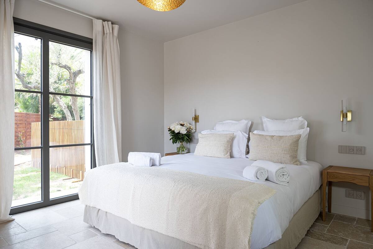 A bedroom with a large bed, white bedding, a bedside table with a lamp, and a view through a window.