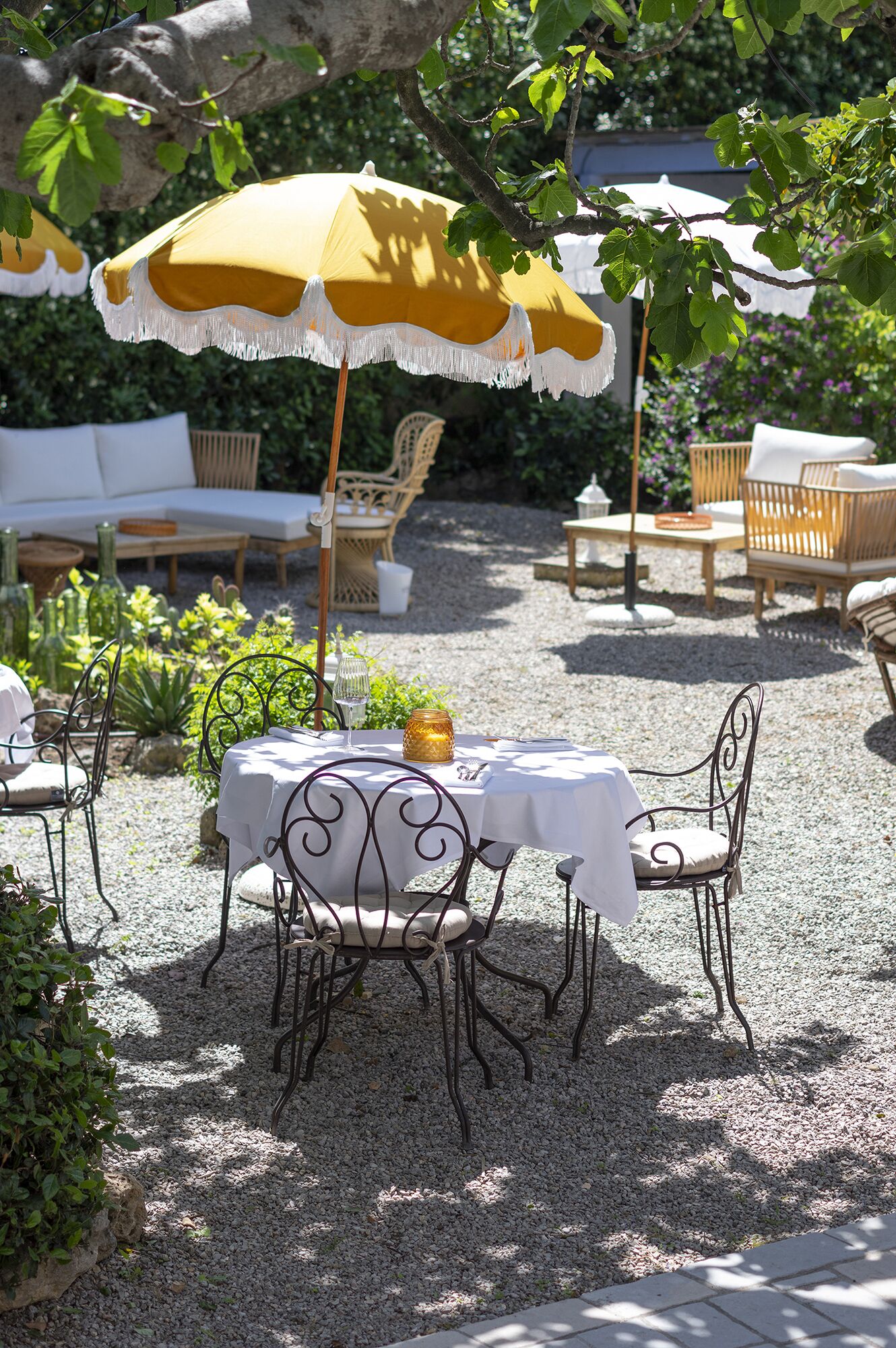 An outdoor patio with a table set for two, surrounded by wicker chairs, under a yellow and white umbrella.