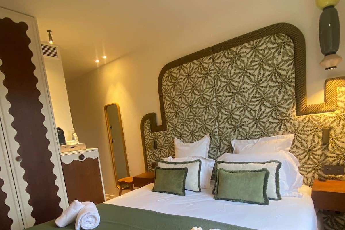 A hotel room with a large bed, green bedspread, and matching pillows. There is a nightstand with a lamp, a mirror, and a small table.