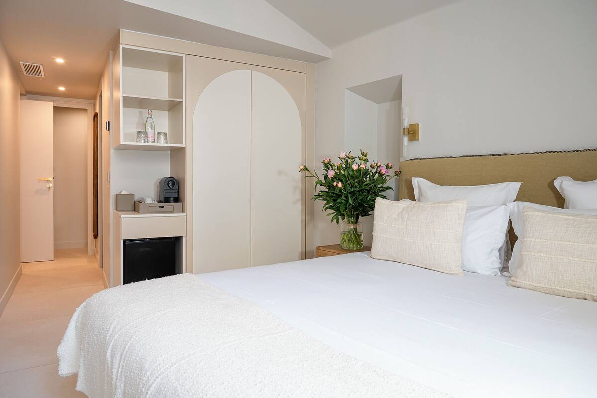 A bedroom with a bed, a nightstand, a bedside table, a wardrobe, and a door leading to another room.