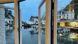 A view through a window of a marina with boats, buildings, and a shop with lights. - Stunning Views of Polperro Harbour from this Waterfront Cottage. Free Parking. (Polperro)
