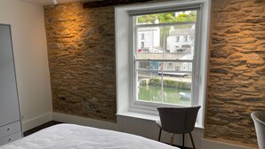 A bedroom with a stone wall, a bed, a chair, and a window overlooking a canal. - Stunning Views of Polperro Harbour from this Waterfront Cottage. Free Parking. (Polperro)