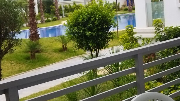 A balcony with a view of a swimming pool, palm trees, and a white building with multiple balconies. - Apartment (Bouznika)