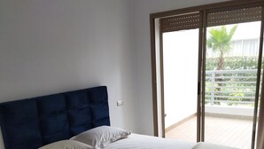 A bedroom with a large bed, a dark headboard, and a view of a balcony with greenery. - Apartment (Bouznika)