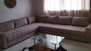 A living room with a beige sectional sofa, a glass coffee table, a cowhide rug, a wall clock, and sheer curtains. - Apartment (Bouznika)