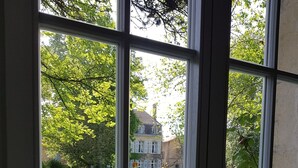 A view through a window with a grid pattern, revealing a grassy area, a pathway, and a building with a chimney in the background. - Orangerie. (Saint-Justin)