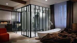 A modern bathroom with a glass enclosure, a freestanding bathtub, and a cozy seating area with a red chair. - Petit Hôtel Confidentiel (Chambery)