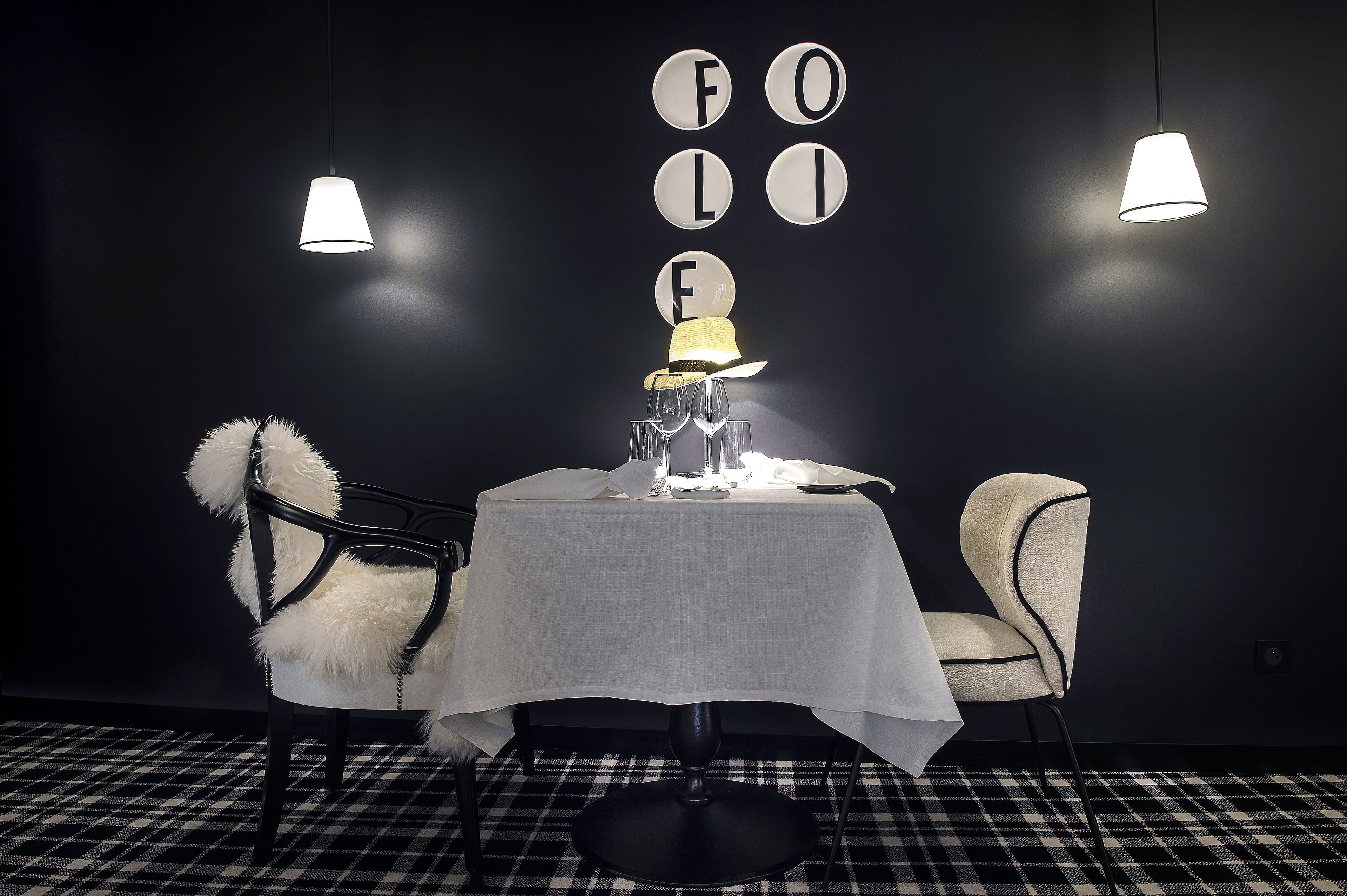 Una mesa vestida con mantel blanco, un sombrero y gafas, sobre un suelo a cuadros blanco y negro, con dos lámparas colgantes.