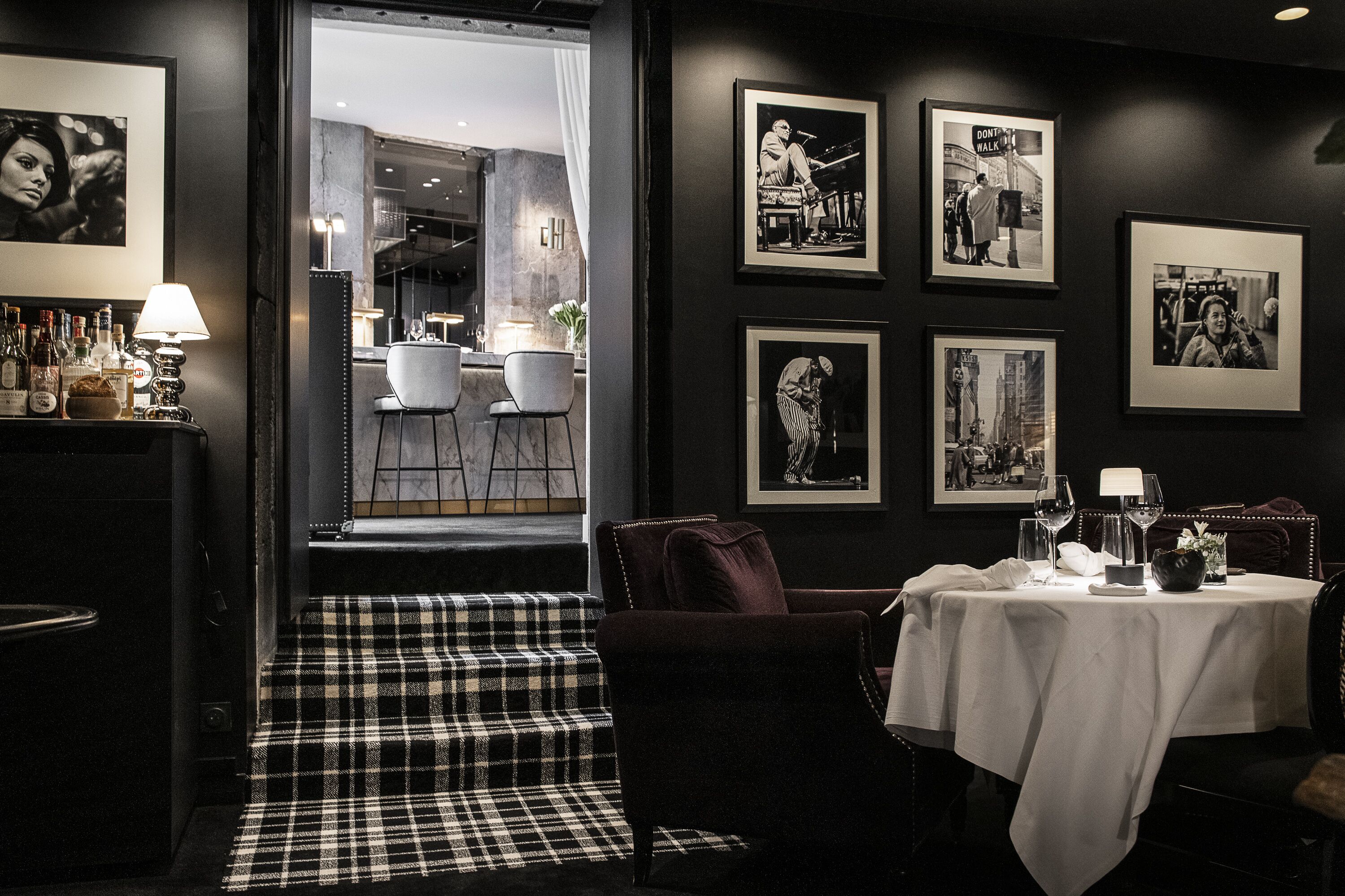 A dining area with a checkered floor, a table set with wine glasses, and framed black and white photographs on the wall.
