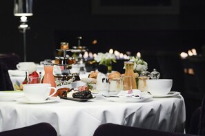 Una mesa lista para el desayuno con diversos platos, entre ellos pasteles y bebidas, en una habitación con poca luz.