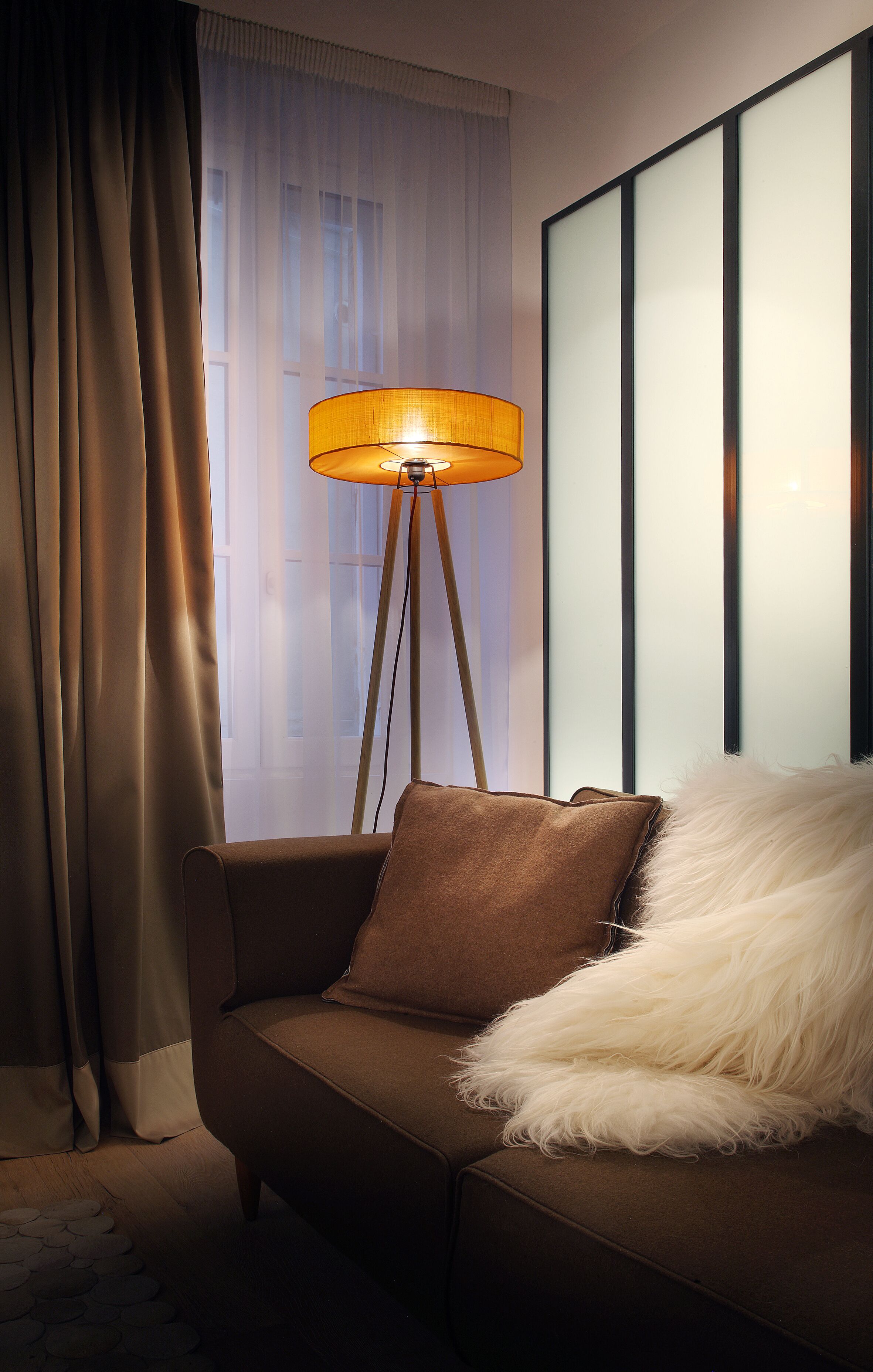 A room with a brown sofa, a fluffy white throw, and a floor lamp with a yellow shade.