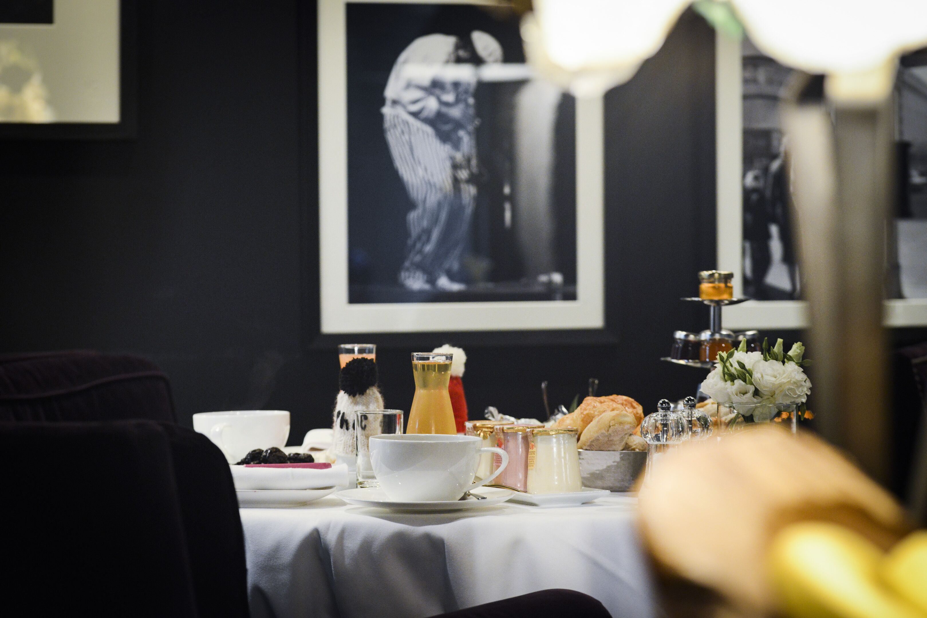 Une table à manger dressée avec une nappe blanche, un verre de jus de fruits, une tasse, une bouteille, un panier à pain et un petit vase avec des fleurs.