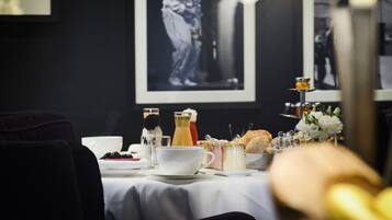 Une table à manger dressée avec une nappe blanche, un verre de jus de fruits, une tasse, une bouteille, un panier à pain et un petit vase avec des fleurs.