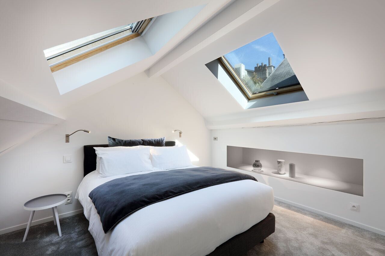 A modern bedroom with a skylight, a bed with white and dark bedding, a bedside table, and a built-in shelf with decorative items.