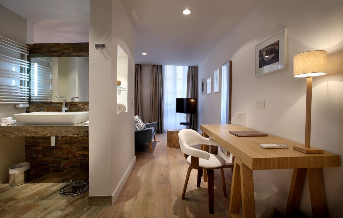 A modern hotel room with a wooden floor, a large sink, a dining table with chairs, a TV, and a lamp.