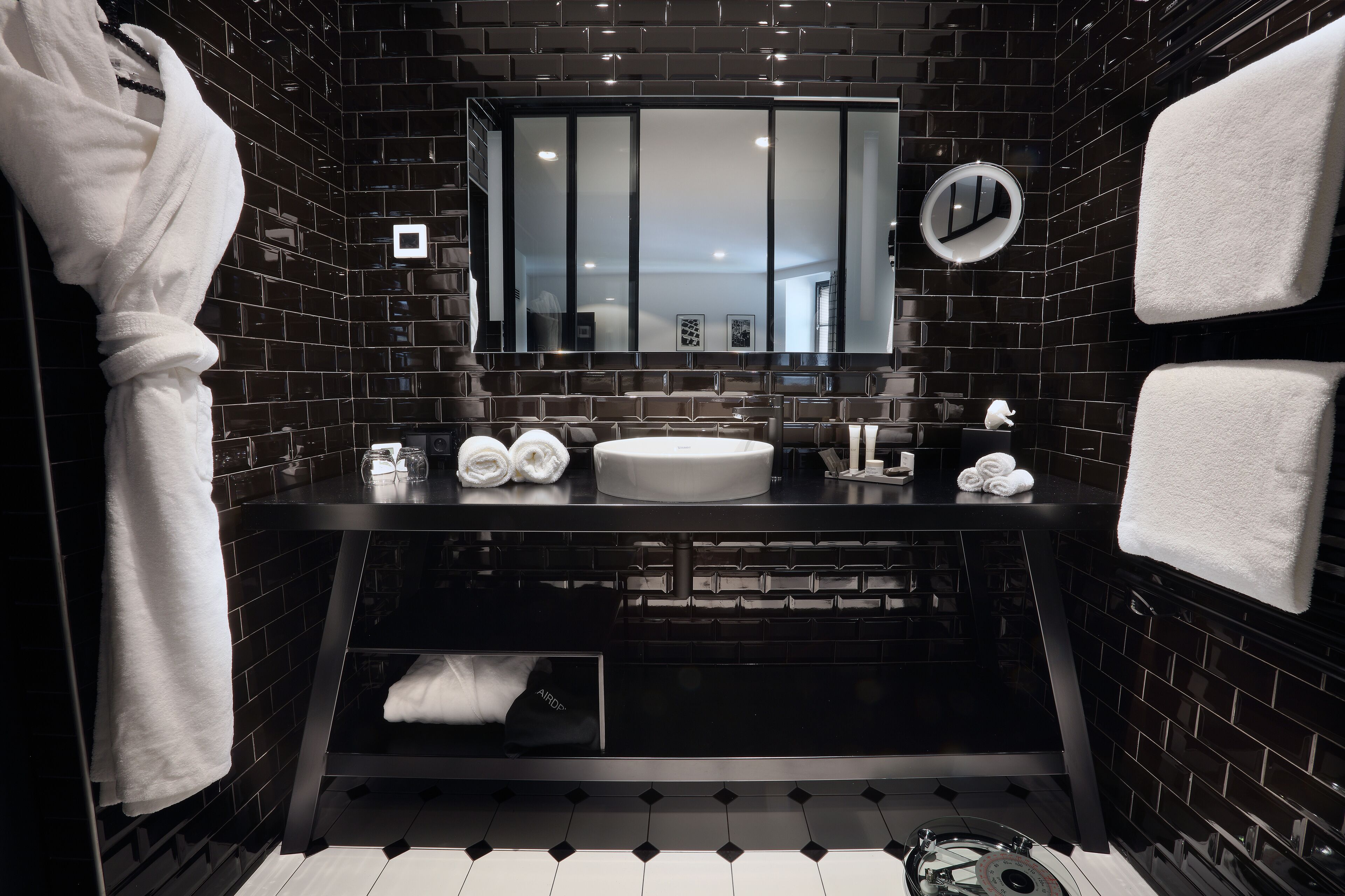 Un baño moderno con paredes de azulejos negros, un espejo grande, un lavabo redondo y toallas ordenadamente dobladas.