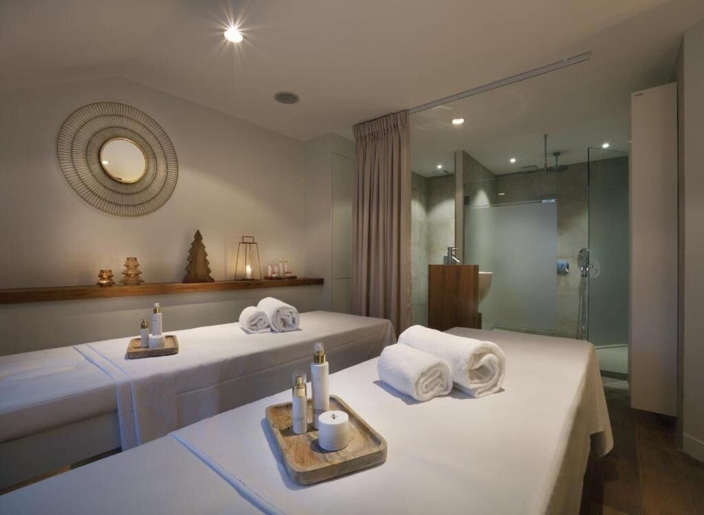 A hotel room with two beds, each with a tray of toiletries and towels. There is a mirror on the wall and a shower area visible through a glass door.