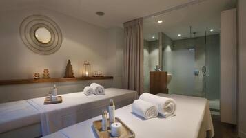 A hotel room with two beds, each with a tray of toiletries and towels. There is a mirror on the wall and a shower area visible through a glass door.