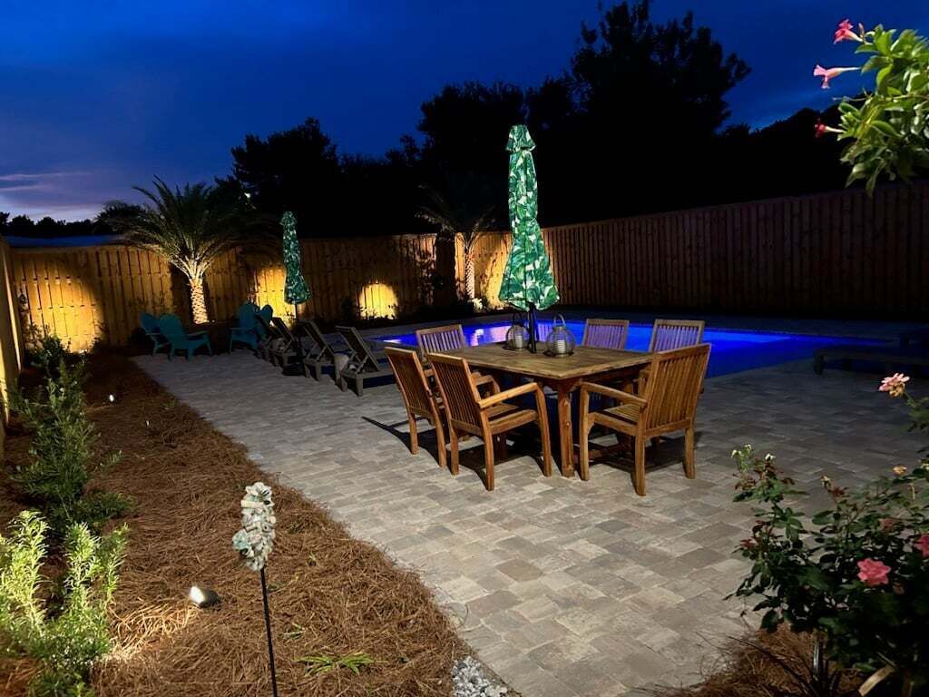 An outdoor patio with a pool, wooden table and chairs, and Christmas lights.