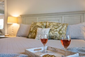 Uma cama com cabeceira de madeira, dois copos de vinho tinto e um tabuleiro com um prato de comida.