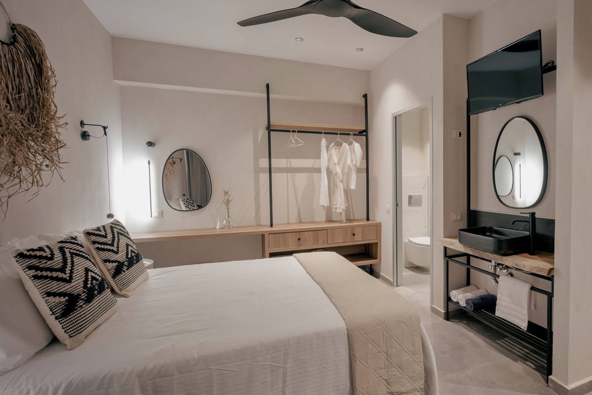 A modern bedroom with a bed, bedside tables, a mirror, a wardrobe, and a bathroom with a sink and mirror.