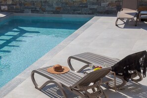Un espace aménagé au bord de la piscine, avec des chaises longues, une table garnie de fruits et un verre de vin, et un mur de pierre en arrière-plan.