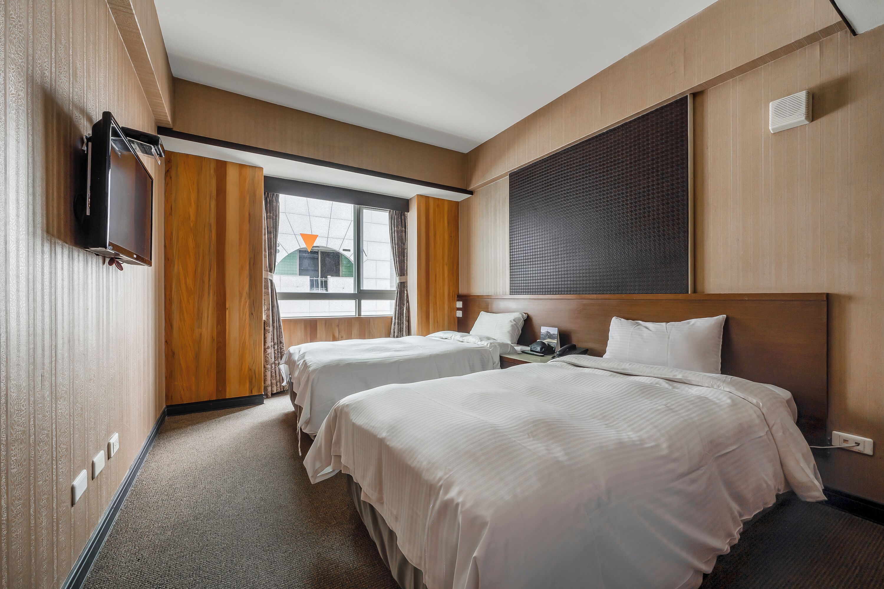 A hotel room with two beds, a TV mounted on the wall, a window with curtains, and a view of a building outside.