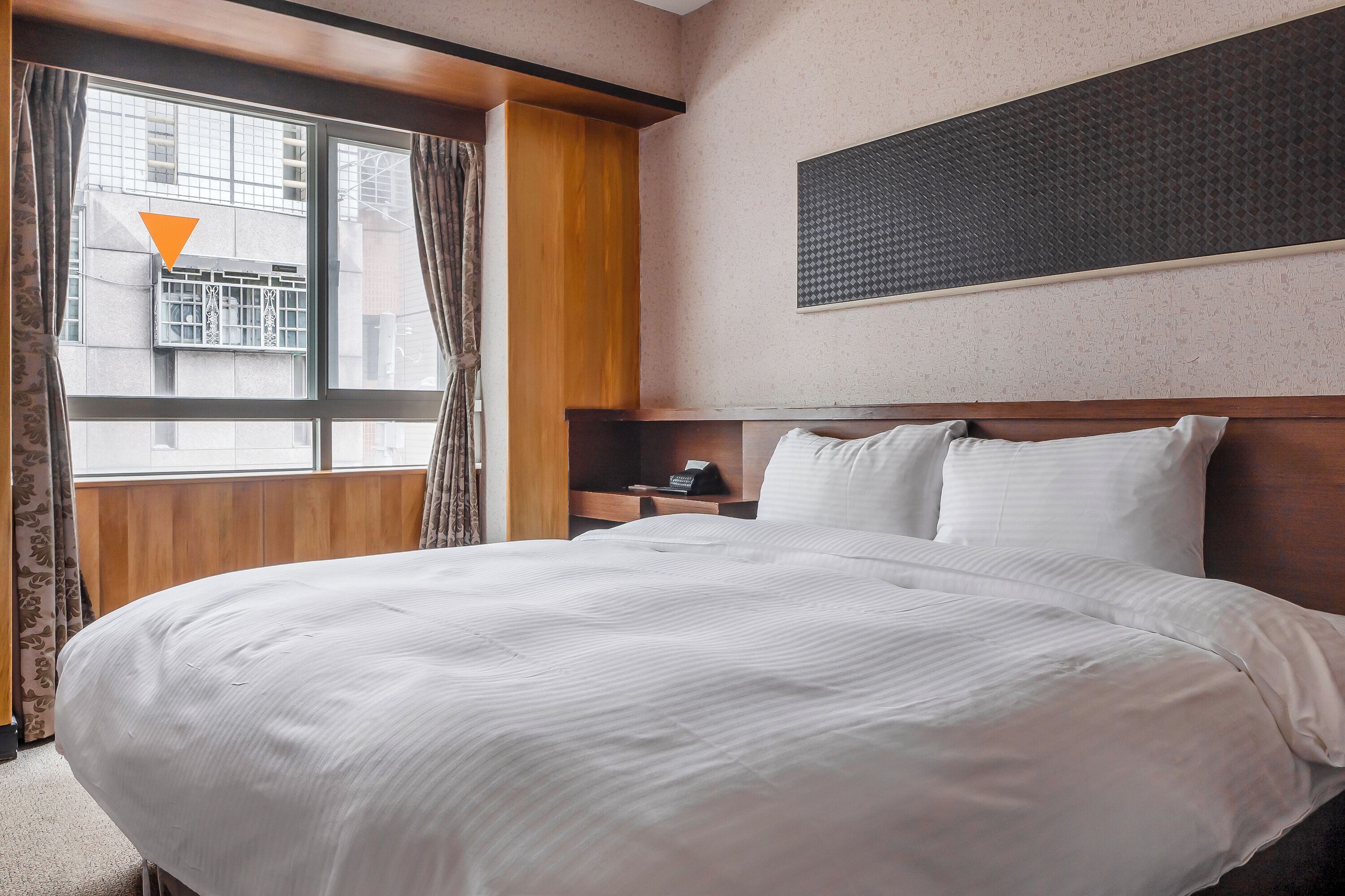 Habitación de hotel con una cama grande, cabecera de madera, mesita de noche con un teléfono y una ventana con cortinas.