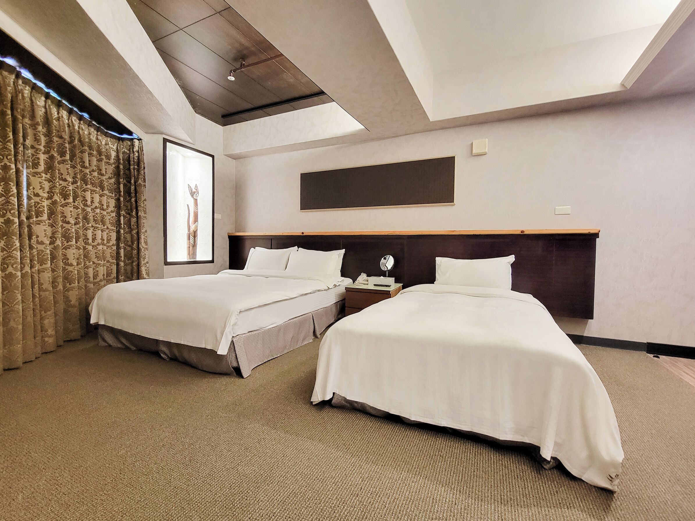 Een hotelkamer met twee bedden, een groot raam met gordijnen, een plafondlamp en een lijst met een afbeelding aan de muur.