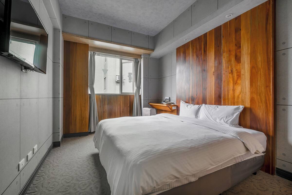 A modern hotel room with a large bed, wooden paneling on the walls, a flat-screen TV mounted on the wall, and a small desk with a chair.