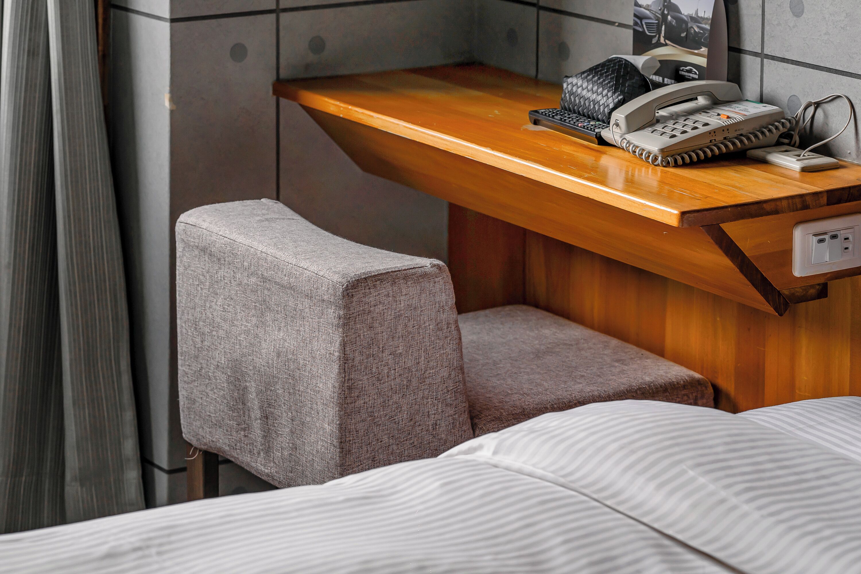 A compact hotel room with a wooden desk, a telephone, and a cushioned bench.