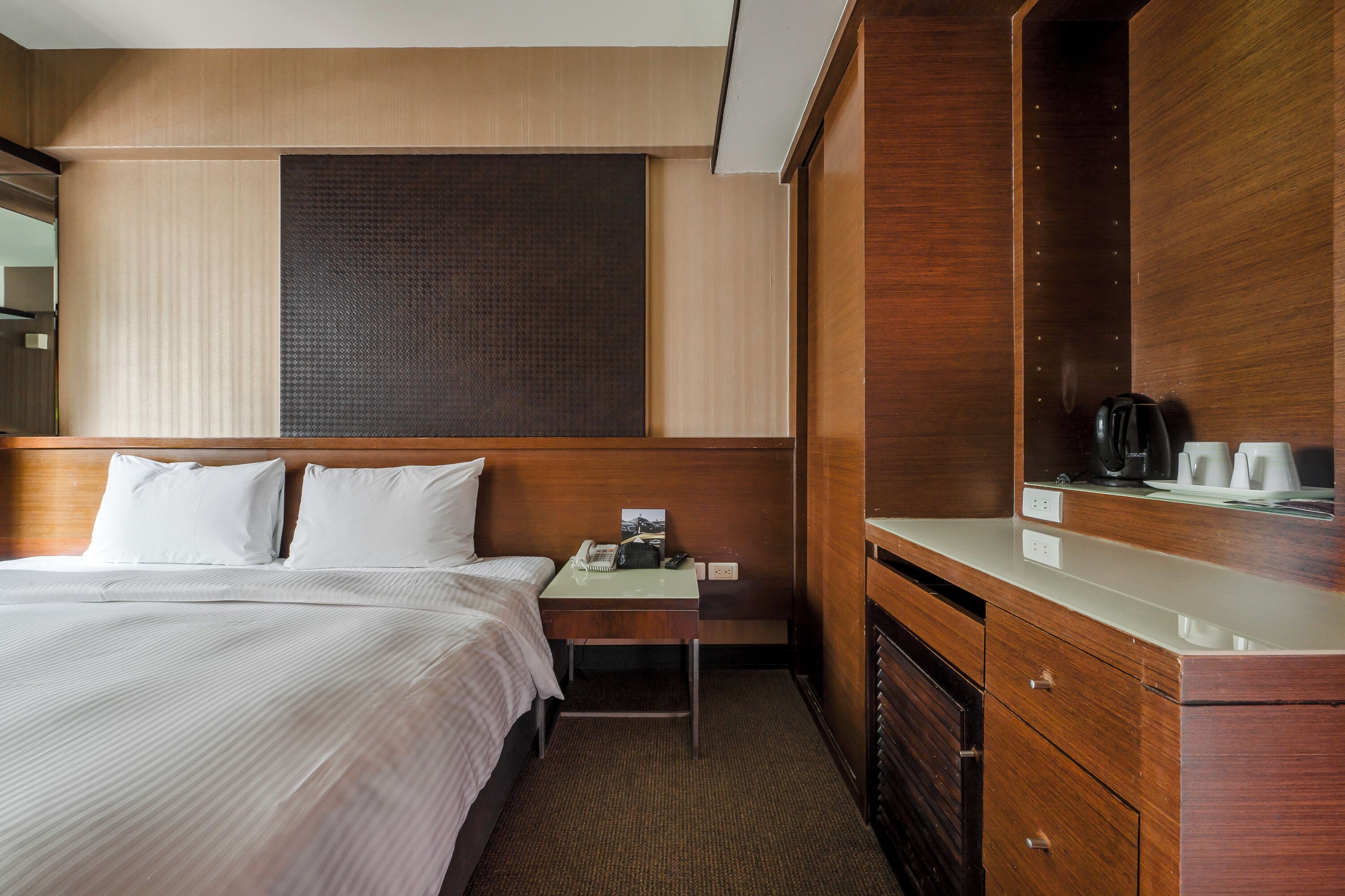 A hotel room with a bed, a bedside table with a phone and a lamp, a wooden dresser, and a large window with blinds.