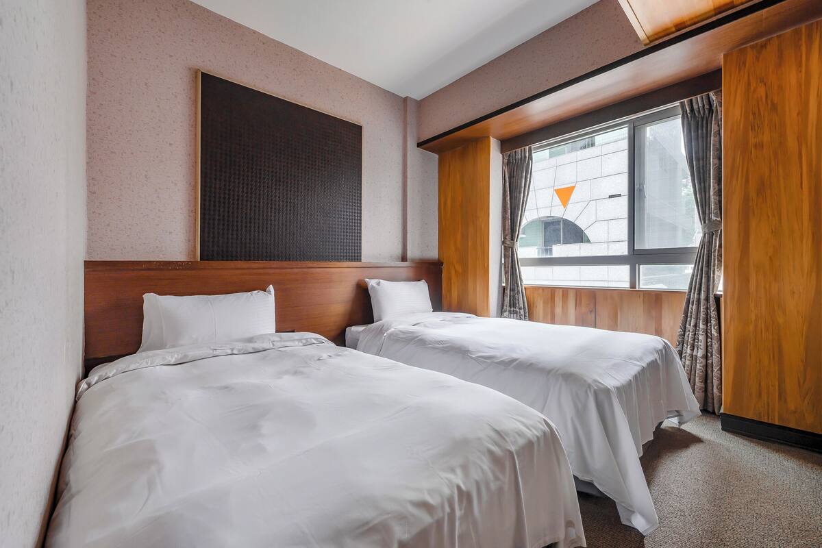 A hotel room with two single beds, a large window with curtains, and a wooden headboard.