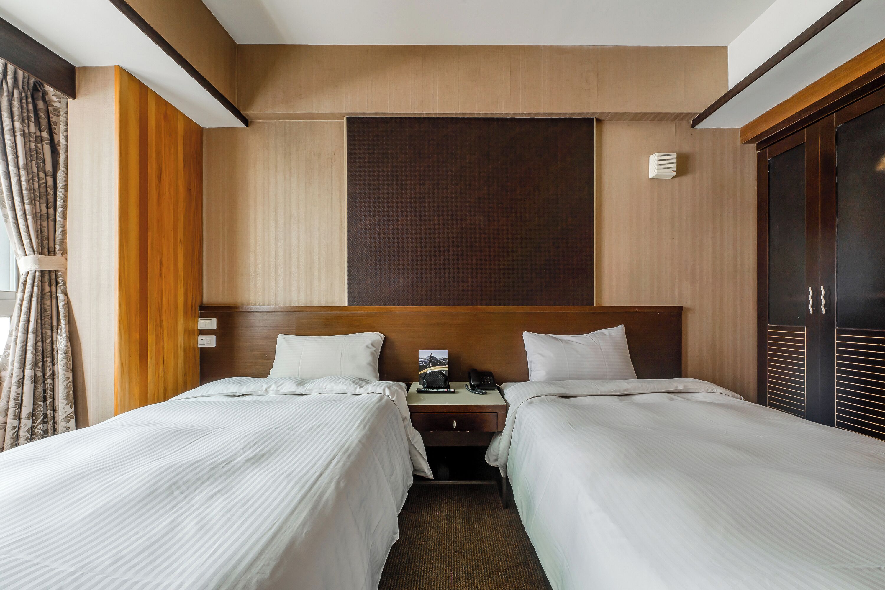 Habitación de hotel con dos camas, cabecera de madera, mesita de noche con teléfono y una ventana grande con cortinas.