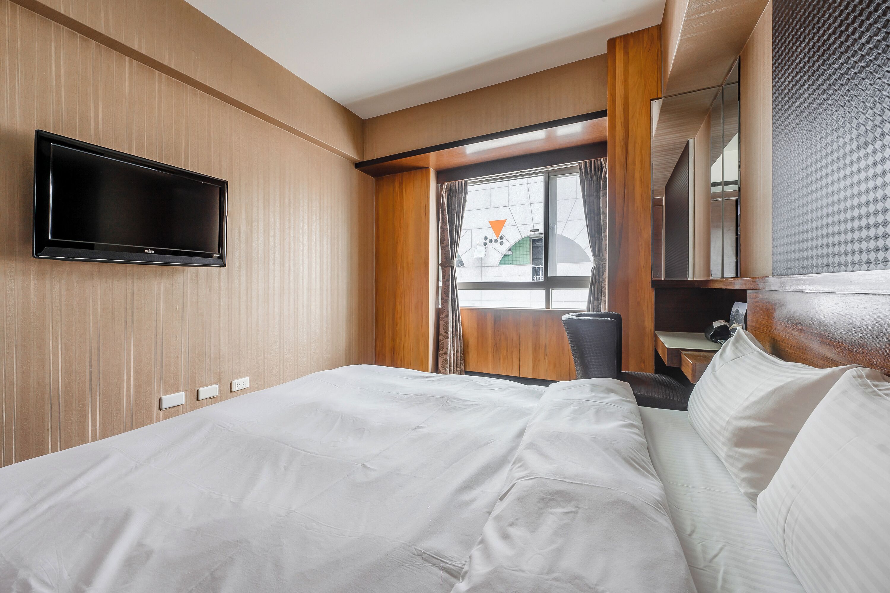 A hotel room with a bed, a flat-screen TV mounted on the wall, a window with curtains, and a desk with a chair.