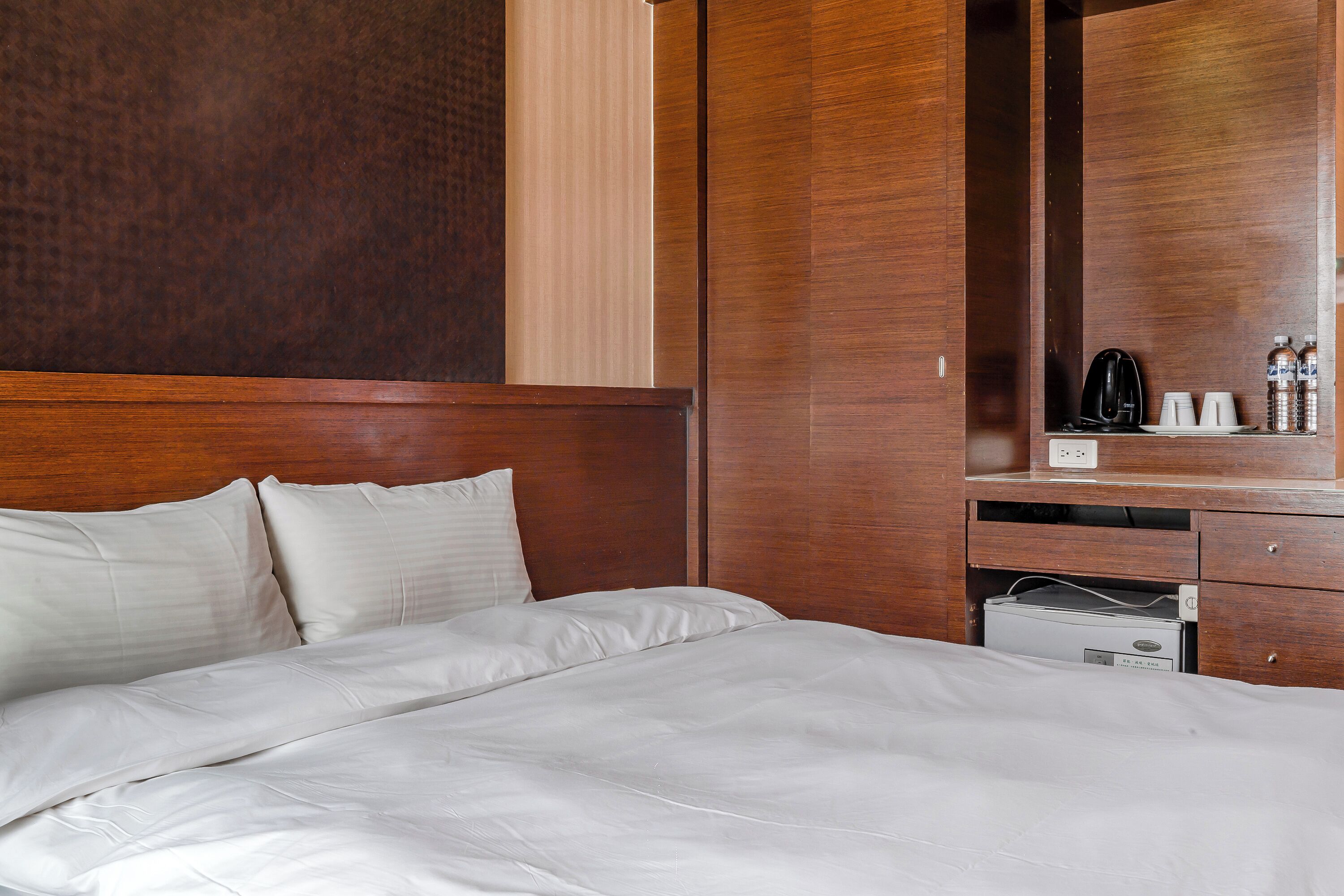 A hotel room with a large bed, wooden headboard, and a built-in wardrobe with a mirror and a small desk area.