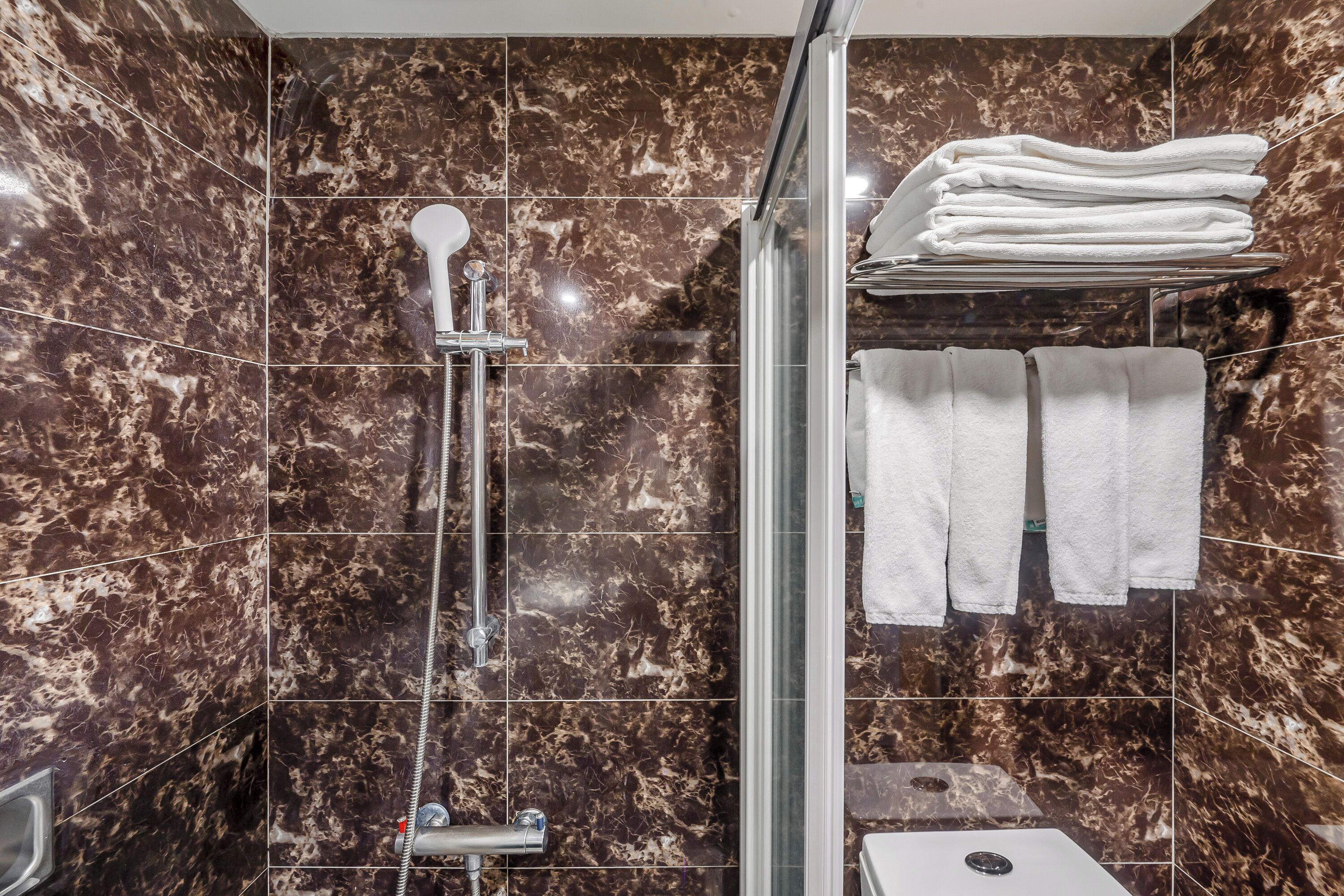 A bathroom with a glass-enclosed shower, marble walls, and a stack of white towels on a shelf.