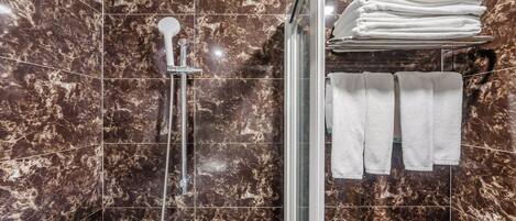 A bathroom with a glass-enclosed shower, marble walls, and a stack of white towels on a shelf.