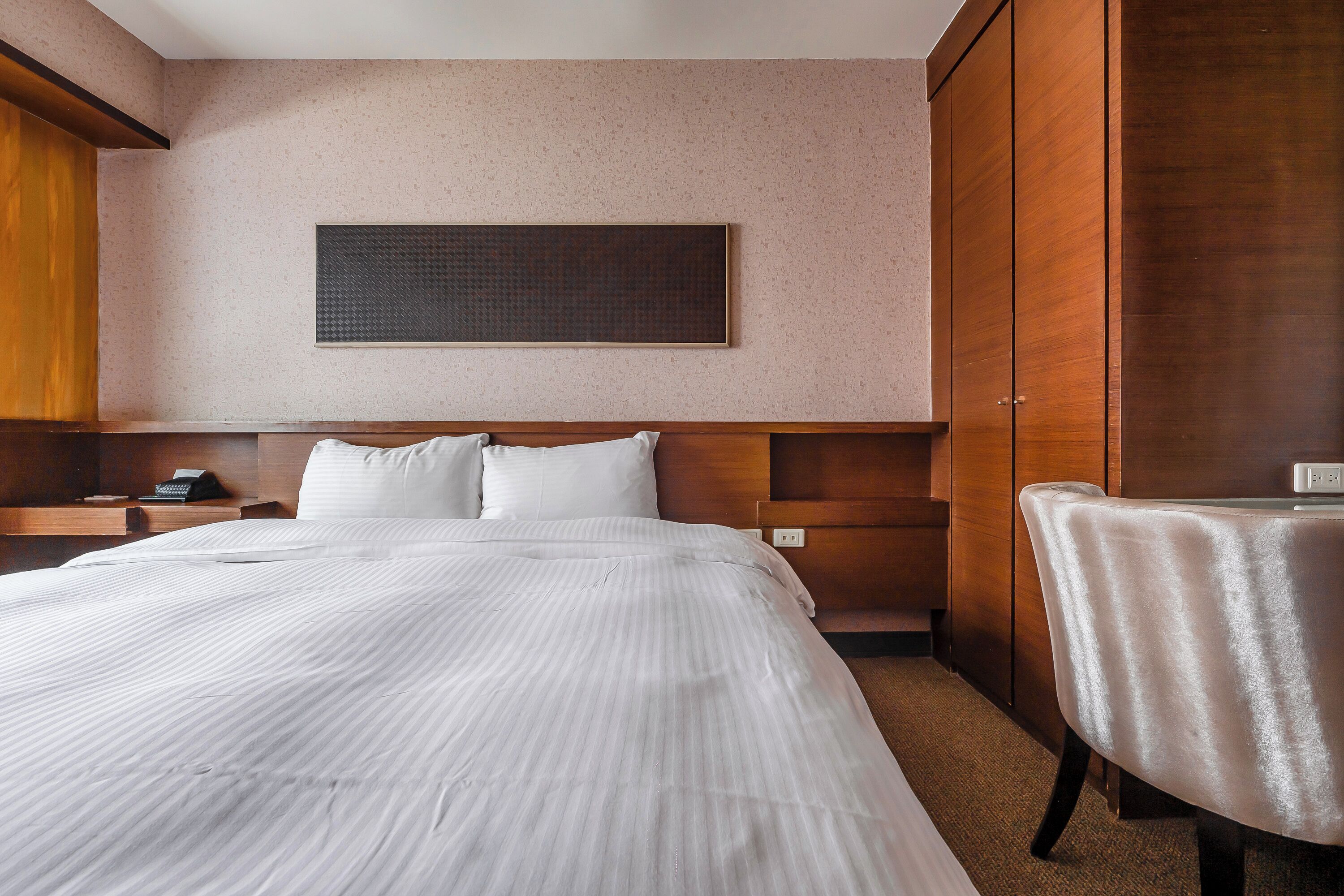 A hotel room with a large bed, a wooden wardrobe, a small table with a cloth, and a wall-mounted artwork.