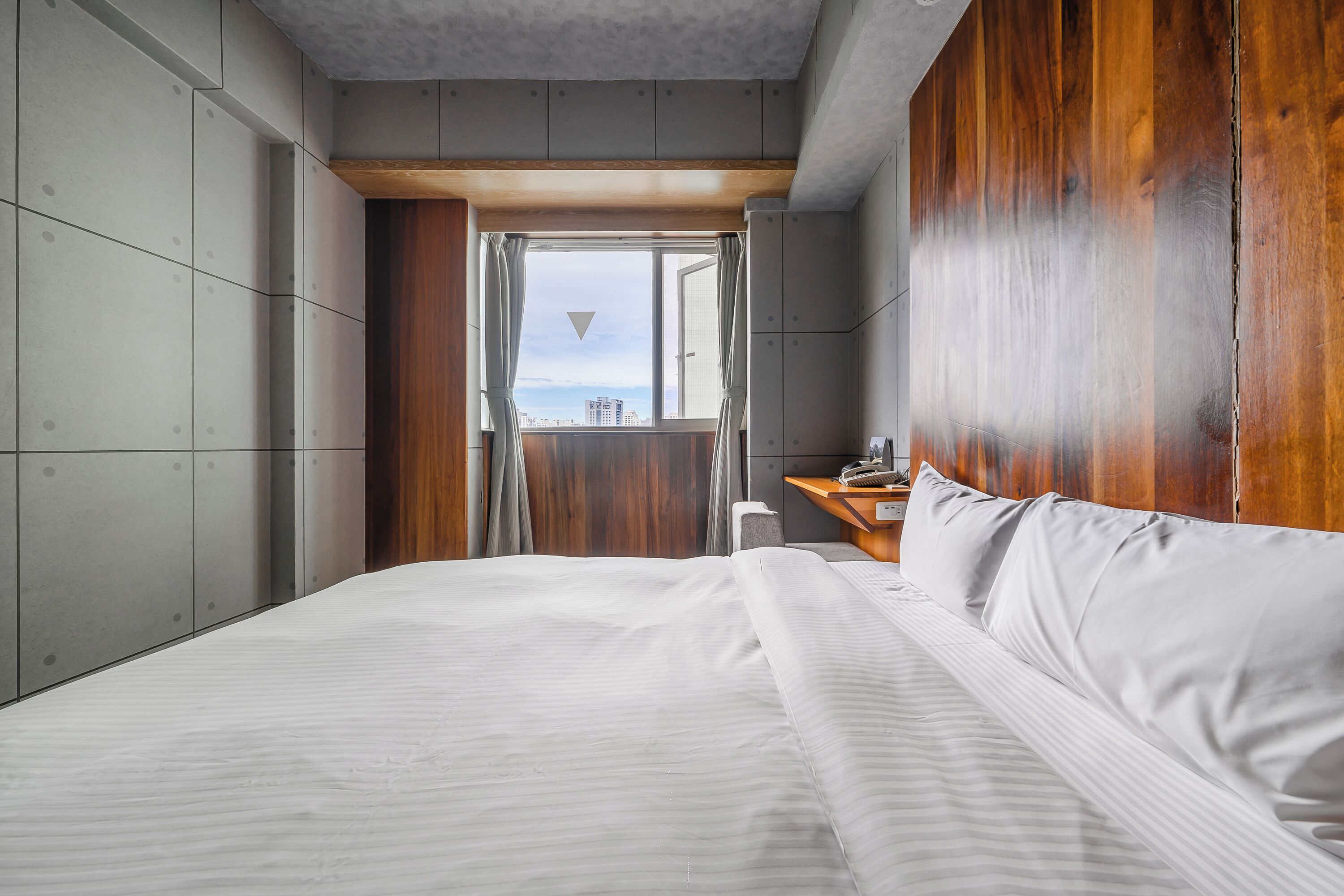 A modern hotel room with a large bed, a wooden headboard, and a view of the city through an open window.