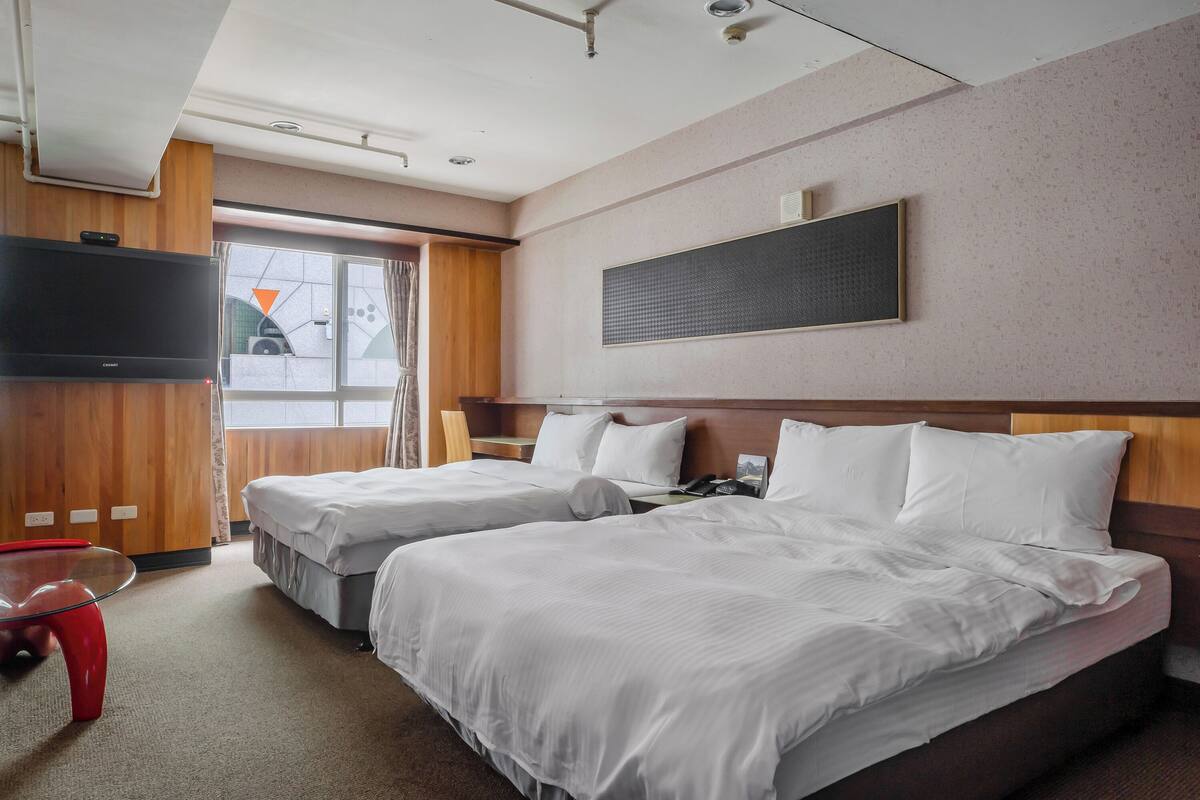 A hotel room with two beds, a TV, a small red table, and a window with curtains.