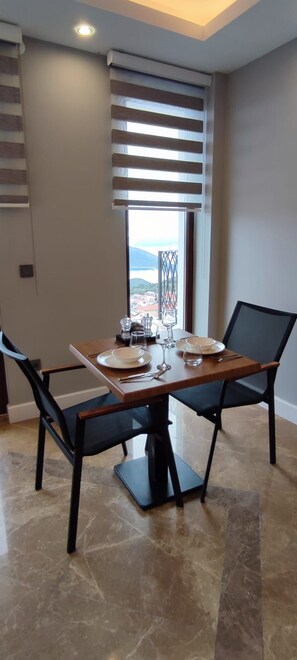 A dining area with a table set for two, a window with blinds, and a view of a bridge and hills. - Arya Deluxe Apart (Kas)