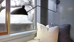 Mukava lukonurkkaus, jossa on ruutukuviollinen koristetyyny, musta pöytävalaisin ja ikkuna, josta avautuu lumipeitteinen maisema.